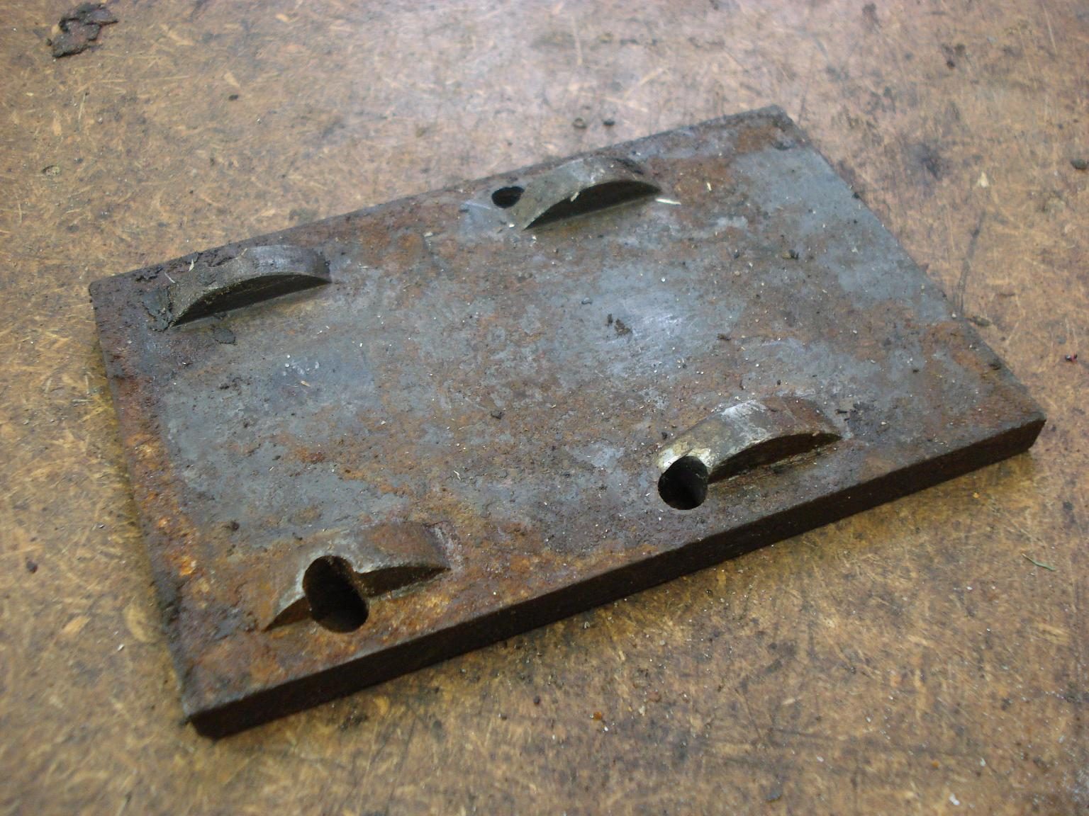 Heavy rectangular machined steel plate with four slotted mounting holes on a workbench.